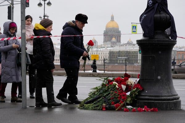 A man in winter clothes places red flowers on a pile of more red flowers.